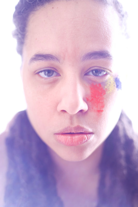 Closeup backlit headshot of a person staring into camera with colored powder beneath their left eye in red, yellow, and blue