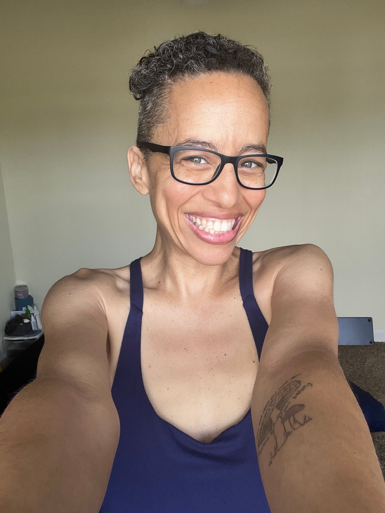 Blue Dulac smiling big, wearing a blue jumpsuit, holding the phone with both hands for a selfie with her mushroom tattoo on left arm visible