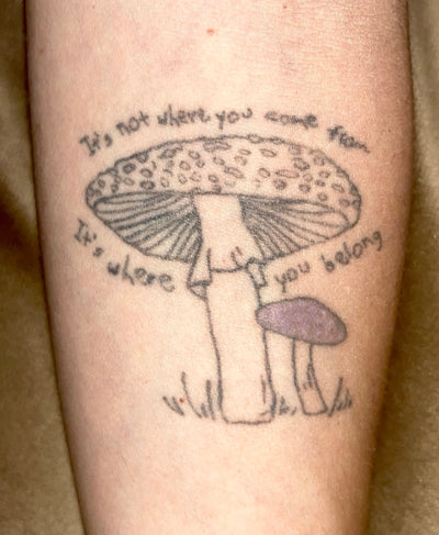 Blue Dulac's arm tattoo of a larger Fly Agaric mushroom in black and white next to a small Viscid Violet Cort mushroom with a purple cap with text that reads It's not where you come from It's where you belong